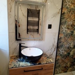 Krzysztof Gardzielik - Nowoczesna łazienka po renowacji: czarna umywalka na drewnianej szafce, lustro z podświetleniem i grzejnik drabinkowy na tle marmurowych płytek.