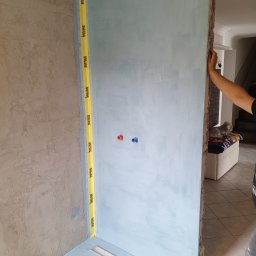 Krzysztof Gardzielik - Przygotowanie ścian w łazience pod płytki: zabezpieczone taśmą krawędzie, nałożona hydroizolacja, widoczne przyłącza wodne. Etap prac przed położeniem płytek.