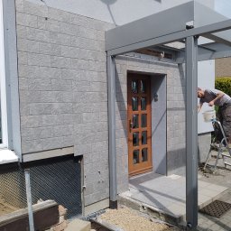 Krzysztof Gardzielik - Montaż elewacji z szarej cegły dekoracyjnej na budynku jednorodzinnym, widoczny fragment ocieplenia i pracownik na drabinie, instalacja zadaszenia nad wejściem.