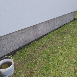 Krzysztof Gardzielik - Wykończona elewacja budynku z szarymi płytkami imitującymi cegłę, widoczna trawa i wiadro z wodą. Detal wykończenia zewnętrznego.