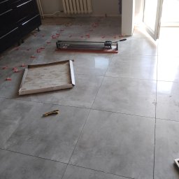 Krzysztof Gardzielik - Szare płytki podłogowe w trakcie układania; widoczna przecinarka, pudełko z płytkami, nożyk i czerwone kliny dystansowe.