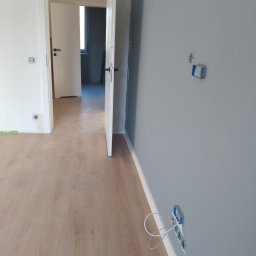 Krzysztof Gardzielik - Jasne panele podłogowe ułożone w jodełkę, szara ściana z puszkami elektrycznymi i otwarte białe drzwi w odnowionym mieszkaniu, widok na sąsiednie pomieszczenie.