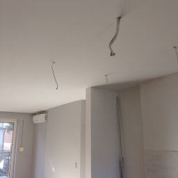 Krzysztof Gardzielik - Wnętrze w trakcie prac wykończeniowych: świeżo pomalowane ściany w odcieniach bieli, widoczne przewody wystające z sufitu, przygotowane do montażu oświetlenia.