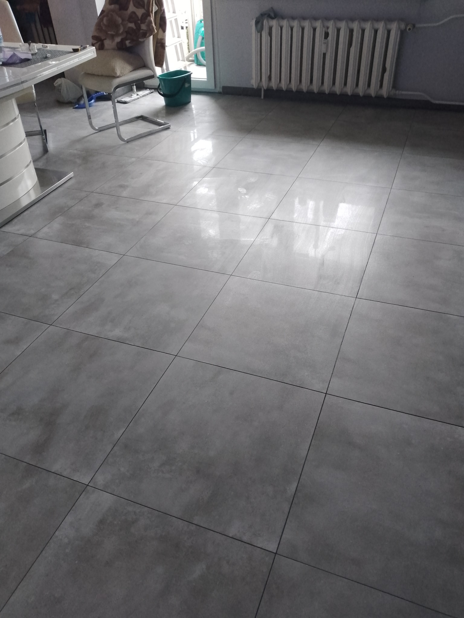 Szare, błyszczące płytki podłogowe ułożone w kwadraty, widoczne w salonie z białym stołem i krzesłem. Grzejnik na ścianie w tle.