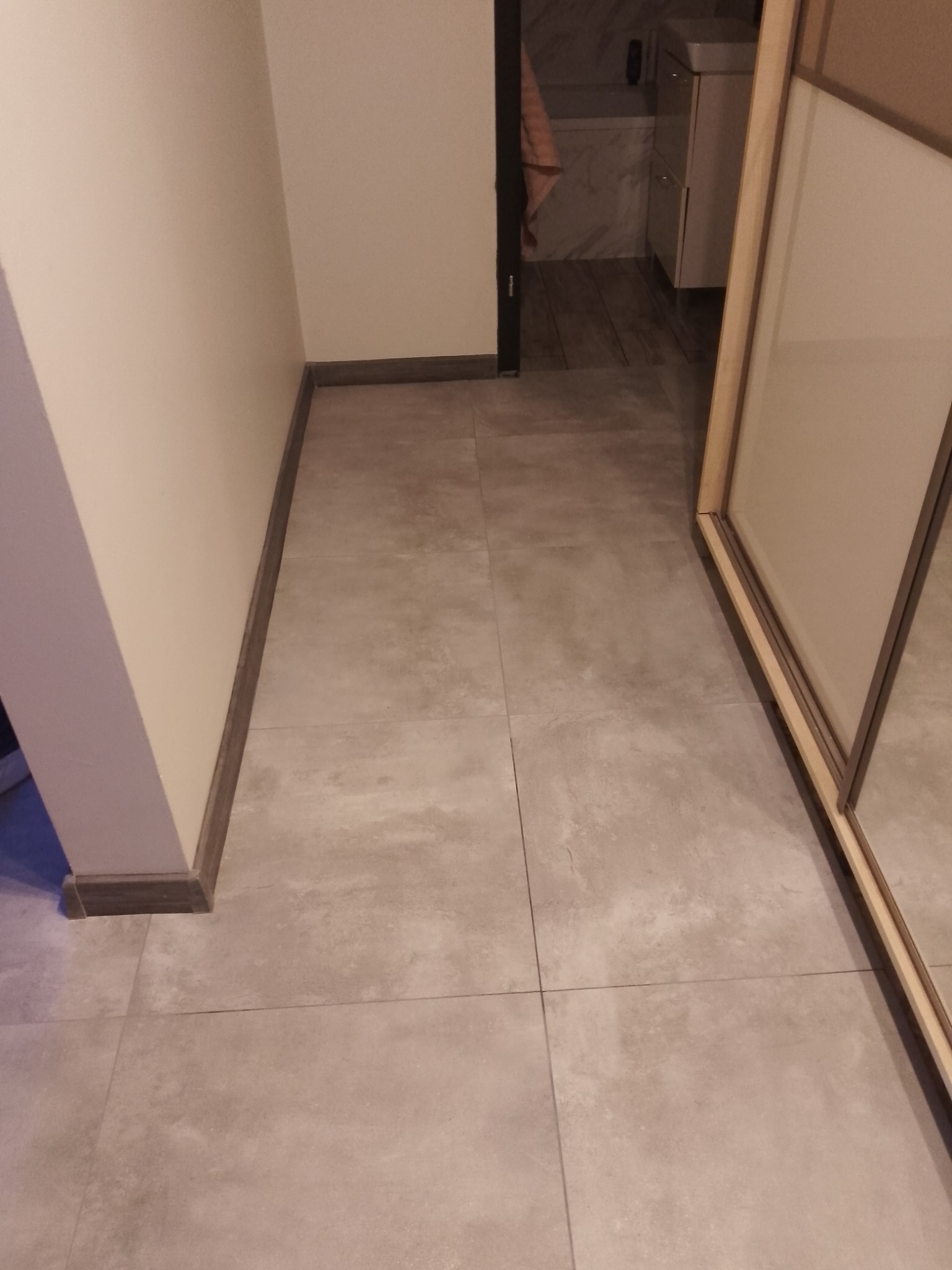 Beżowe płytki podłogowe w korytarzu, z widoczną listwą przypodłogową i fragmentem szafy przesuwnej. W tle widoczna łazienka.