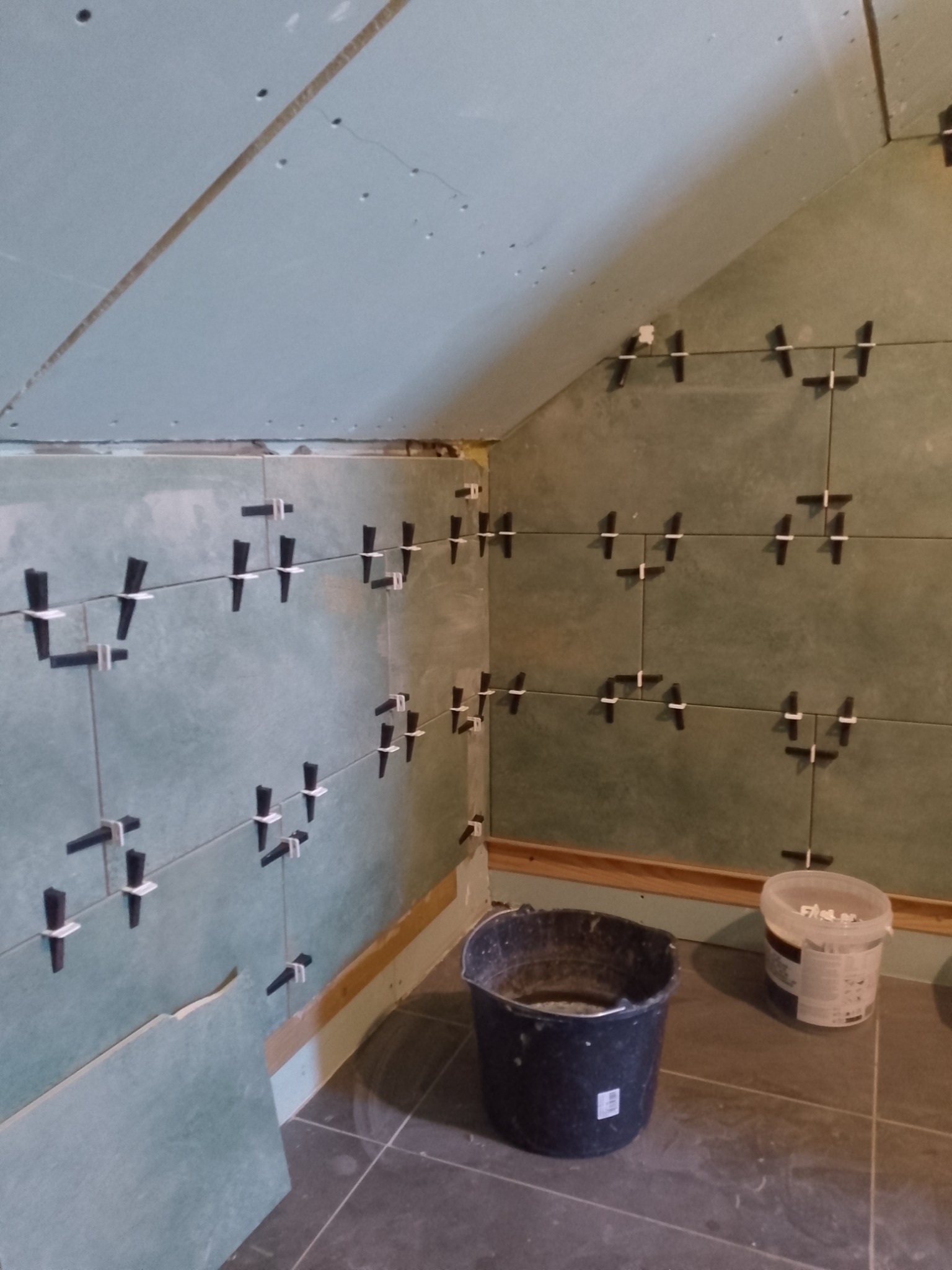 Układanie płytek w łazience na poddaszu z użyciem klipsów poziomujących. Widoczne wiaderka z zaprawą i fragment płyty kartonowo-gipsowej.