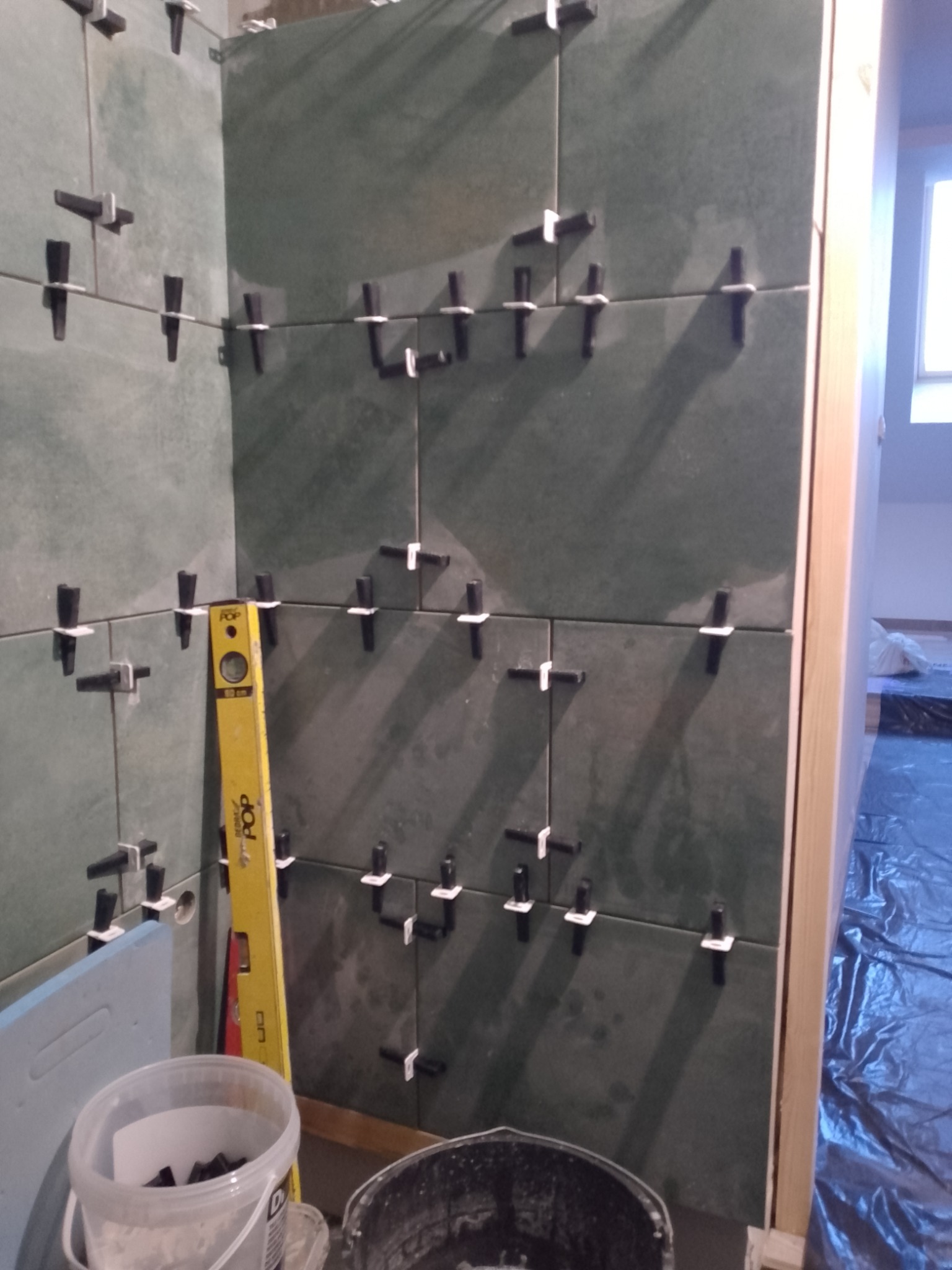 Proces układania ciemnozielonych płytek w rogu łazienki, z użyciem poziomicy i klipsów do wyrównywania, widoczne narzędzia i wiaderka. Prace w toku.