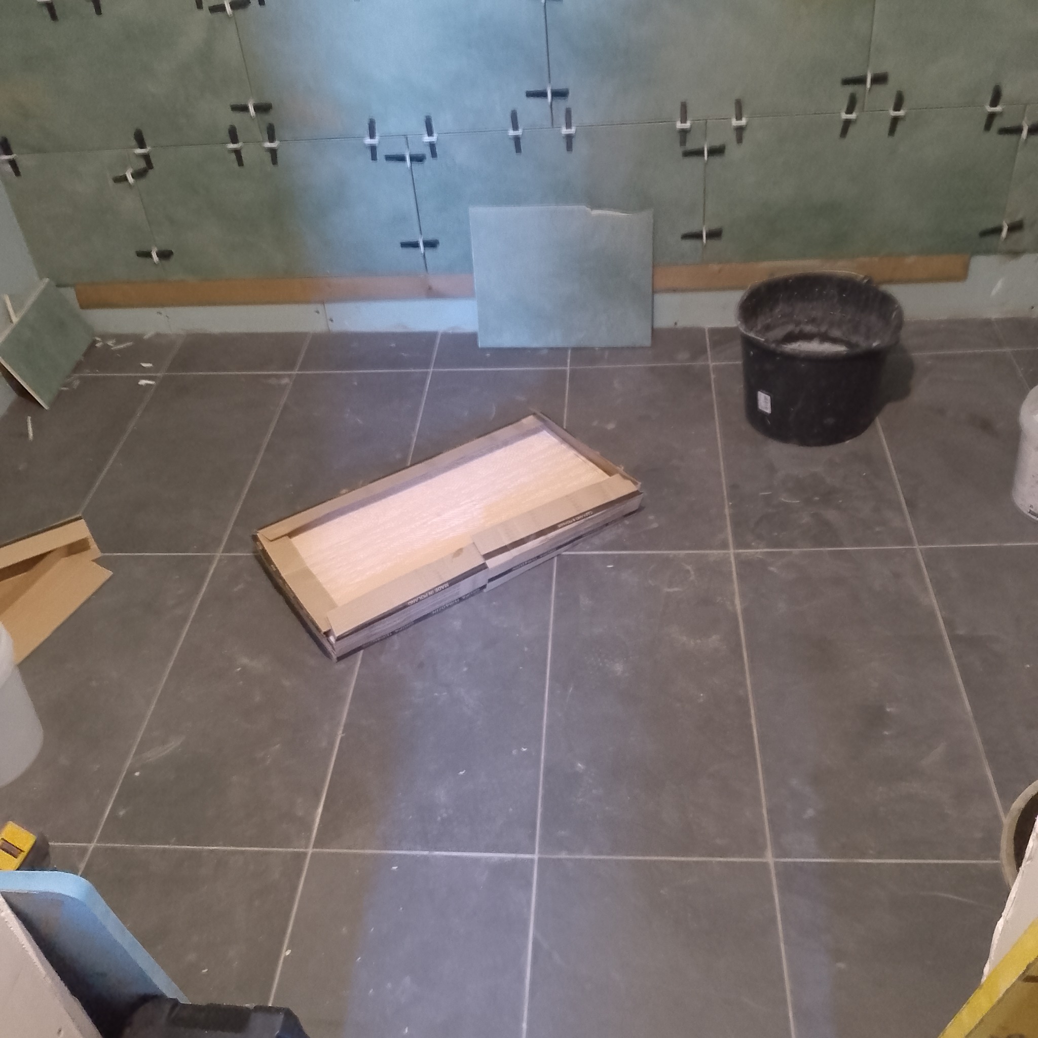 Etap prac wykończeniowych w łazience: montaż płytek na ścianie z użyciem krzyżyków dystansowych, na podłodze karton z panelami i wiadro z zaprawą.