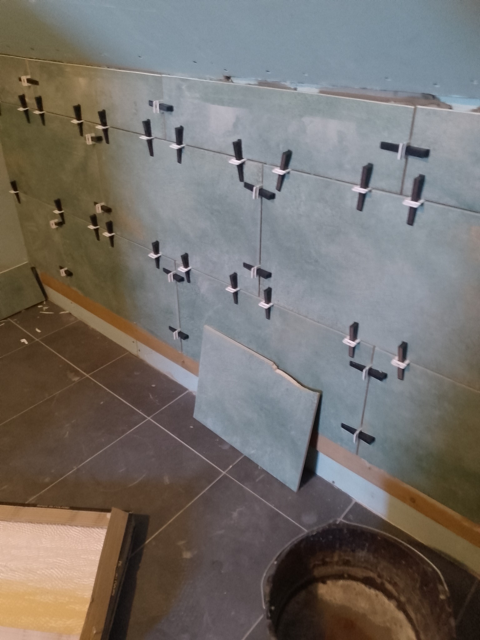 Ściana w trakcie układania płytek ceramicznych w łazience. Widoczne krzyżyki dystansowe i fragment podłogi z ciemnych płytek.