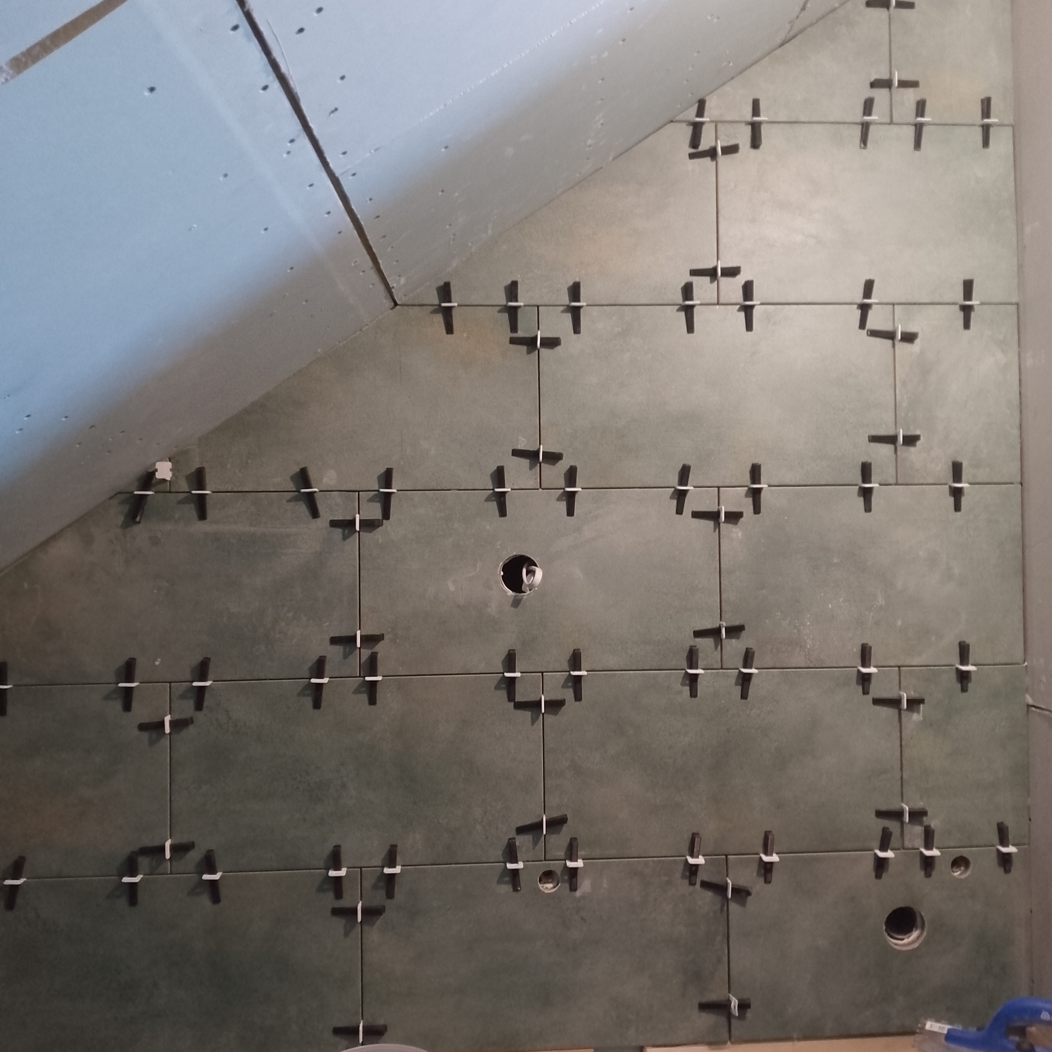 Ściana w skosie wyłożona szarymi płytkami ceramicznymi z użyciem krzyżyków dystansowych. Widoczne otwory na instalacje. Płytki ułożone równo z zachowaniem spoin.