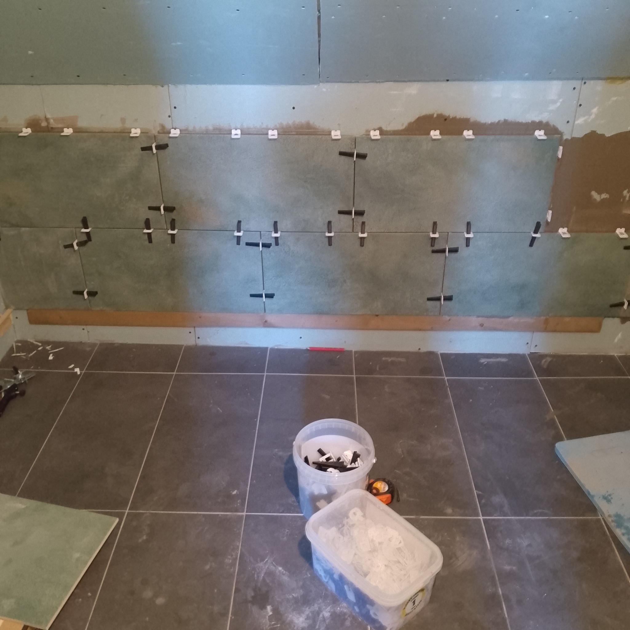 Ściana w trakcie układania płytek, widoczne krzyżyki dystansowe, poziomica, wiaderka z zaprawą i płytki podłogowe. Prace wykończeniowe w łazience.