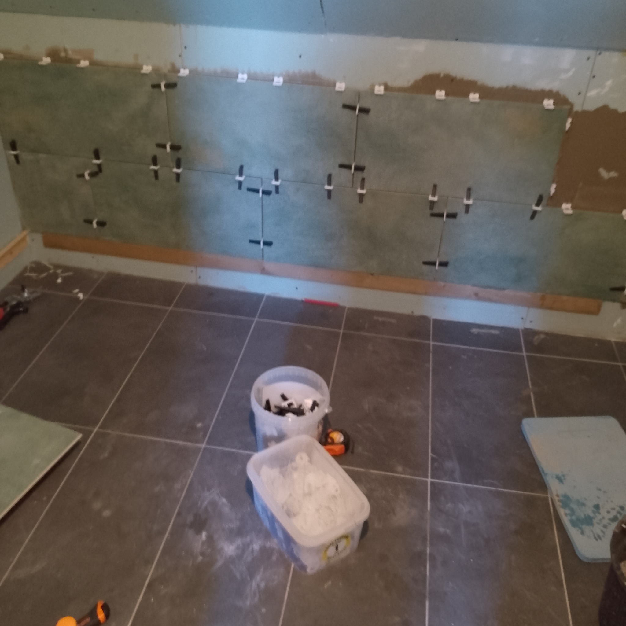 Układanie ciemnych płytek na ścianie w łazience. Widoczne krzyżyki dystansowe, wiaderka z klejem i narzędzia. Etap prac wykończeniowych w trakcie remontu.