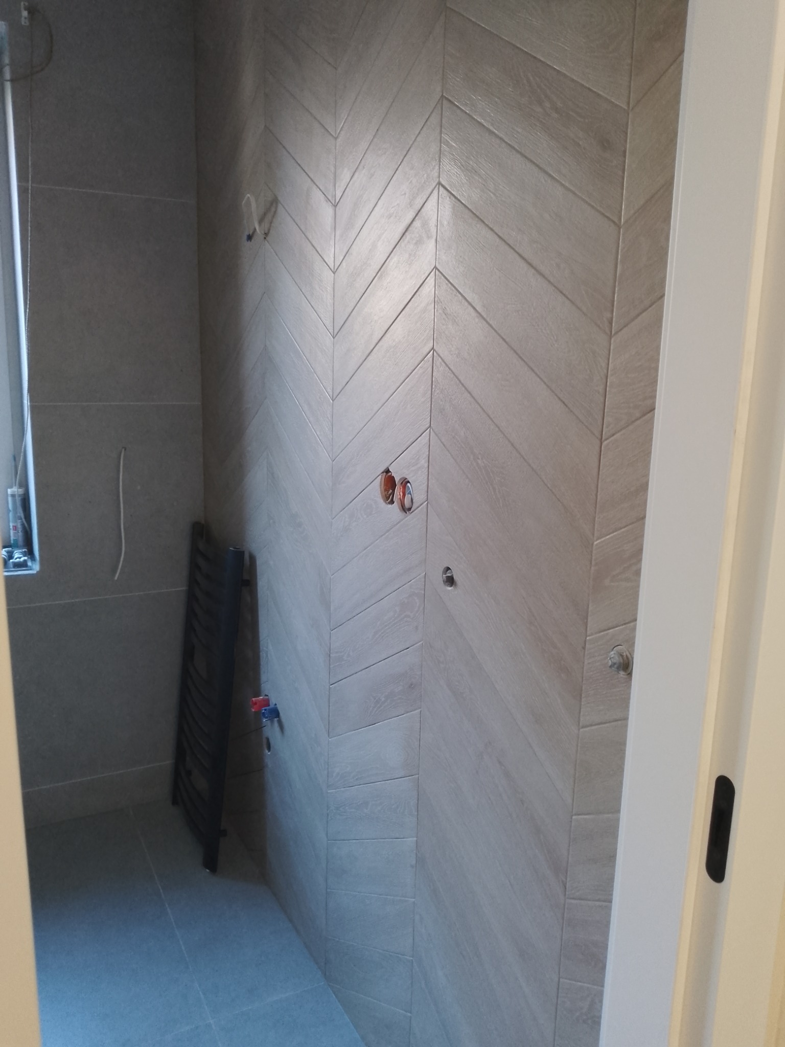 Wykończona łazienka z szarymi płytkami ułożonymi w jodełkę na ścianie, widoczny grzejnik drabinkowy i fragment drzwi. Minimalistyczny design.