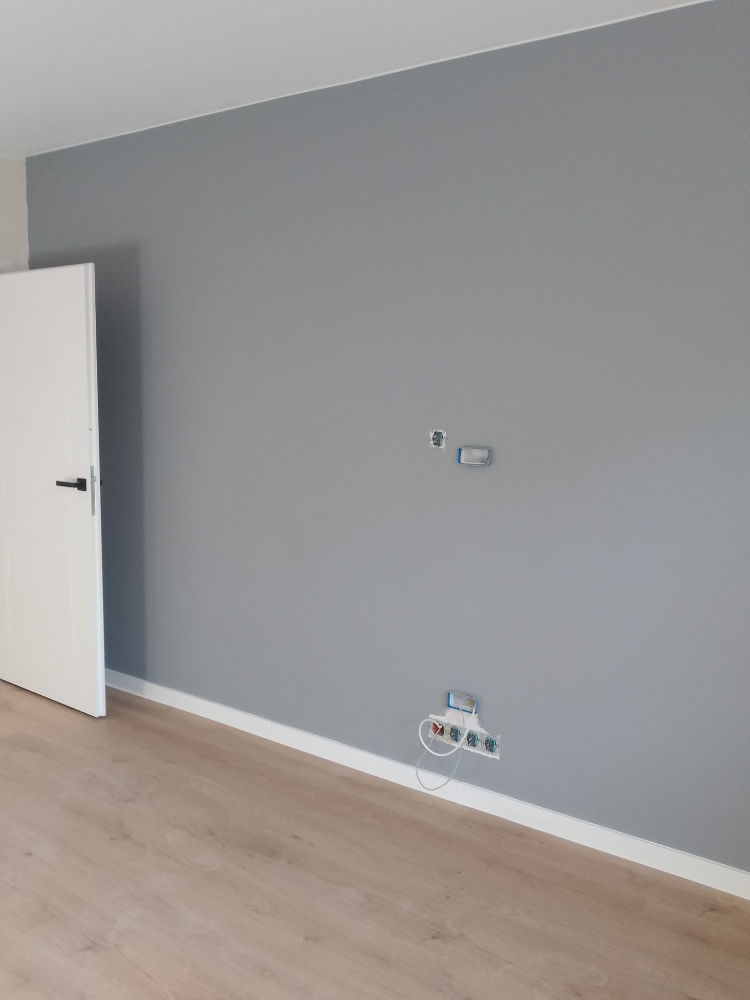 Ściana pomalowana na szaro z gniazdkami elektrycznymi, biała listwa przypodłogowa i jasne panele podłogowe. Widoczne białe drzwi z czarną klamką.