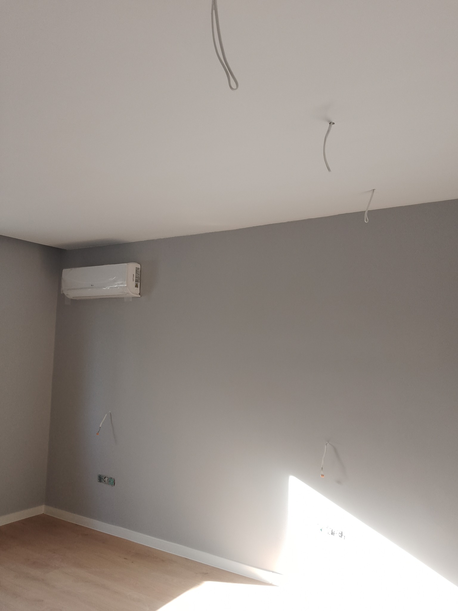 Pokój po malowaniu ścian na szaro, z zamontowanym klimatyzatorem, widoczne wystające kable i gniazdko elektryczne. Na podłodze panele.