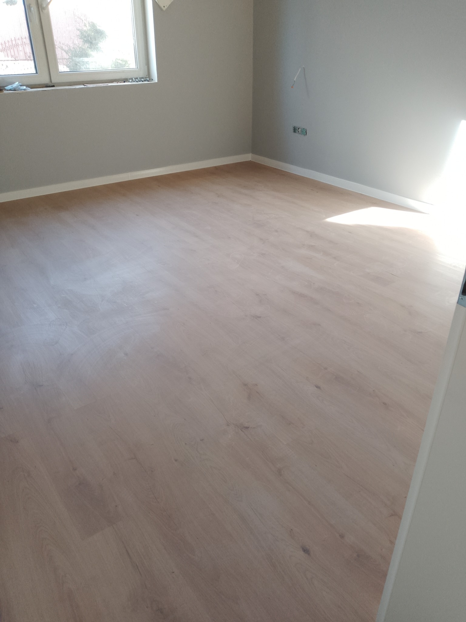 Jasne panele podłogowe ułożone w pokoju z szarymi ścianami i białymi listwami przypodłogowymi, widok na okno i fragment ściany z gniazdkiem.