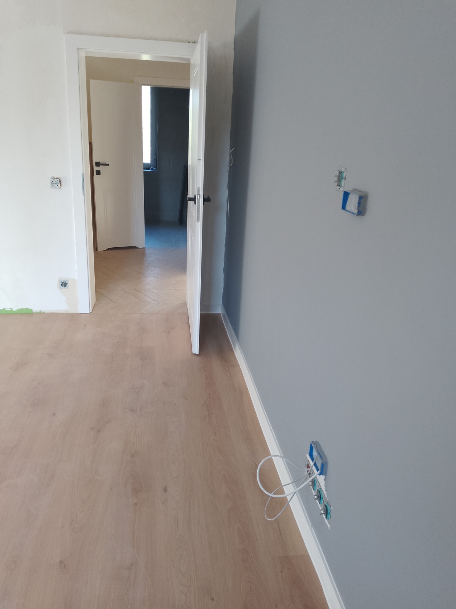 Jasne panele podłogowe ułożone w jodełkę, szara ściana z puszkami elektrycznymi i otwarte białe drzwi w odnowionym mieszkaniu, widok na sąsiednie pomieszczenie.