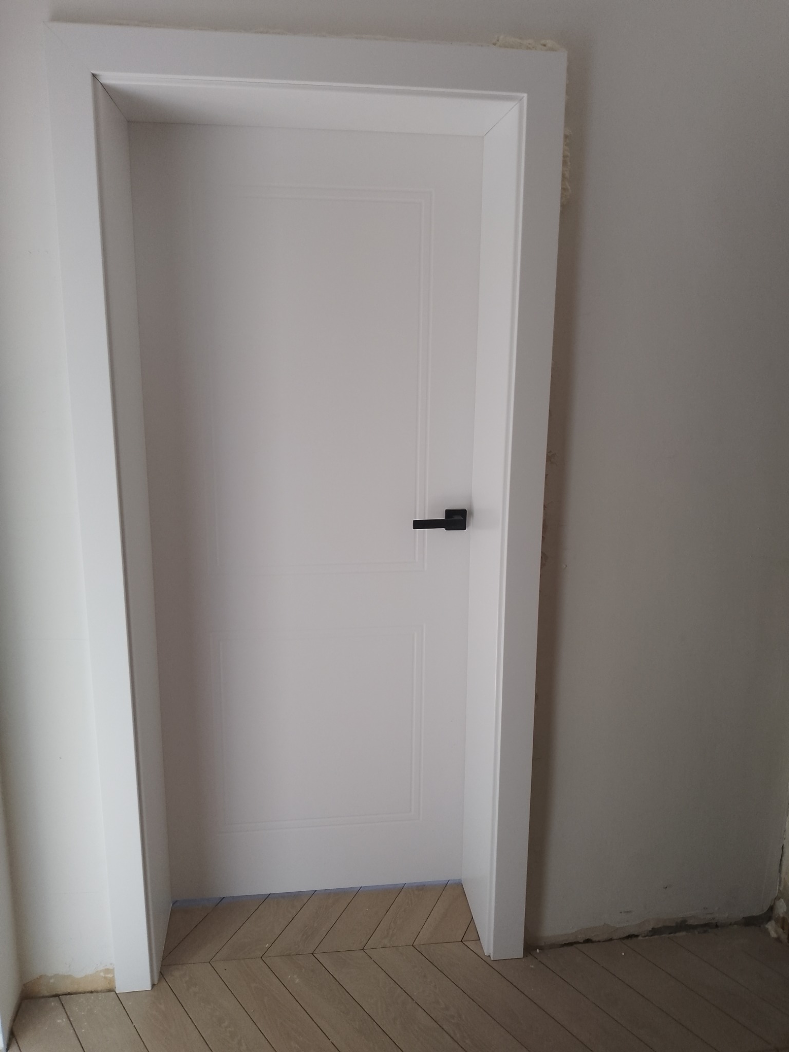 Białe, panelowe drzwi z czarną klamką, osadzone w białej ościeżnicy, na tle jasnych ścian i podłogi ułożonej w jodełkę. Widoczne ślady prac wykończeniowych.