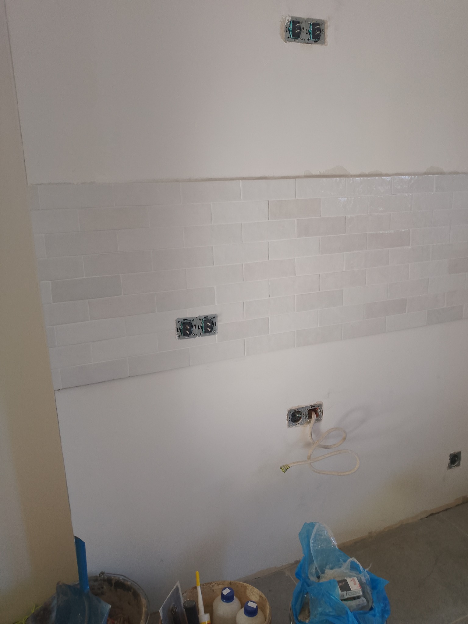 Ściana wyłożona białymi, błyszczącymi płytkami w stylu cegiełki, z widocznymi puszkami elektrycznymi. Częściowo wykończona powierzchnia, w tle narzędzia.