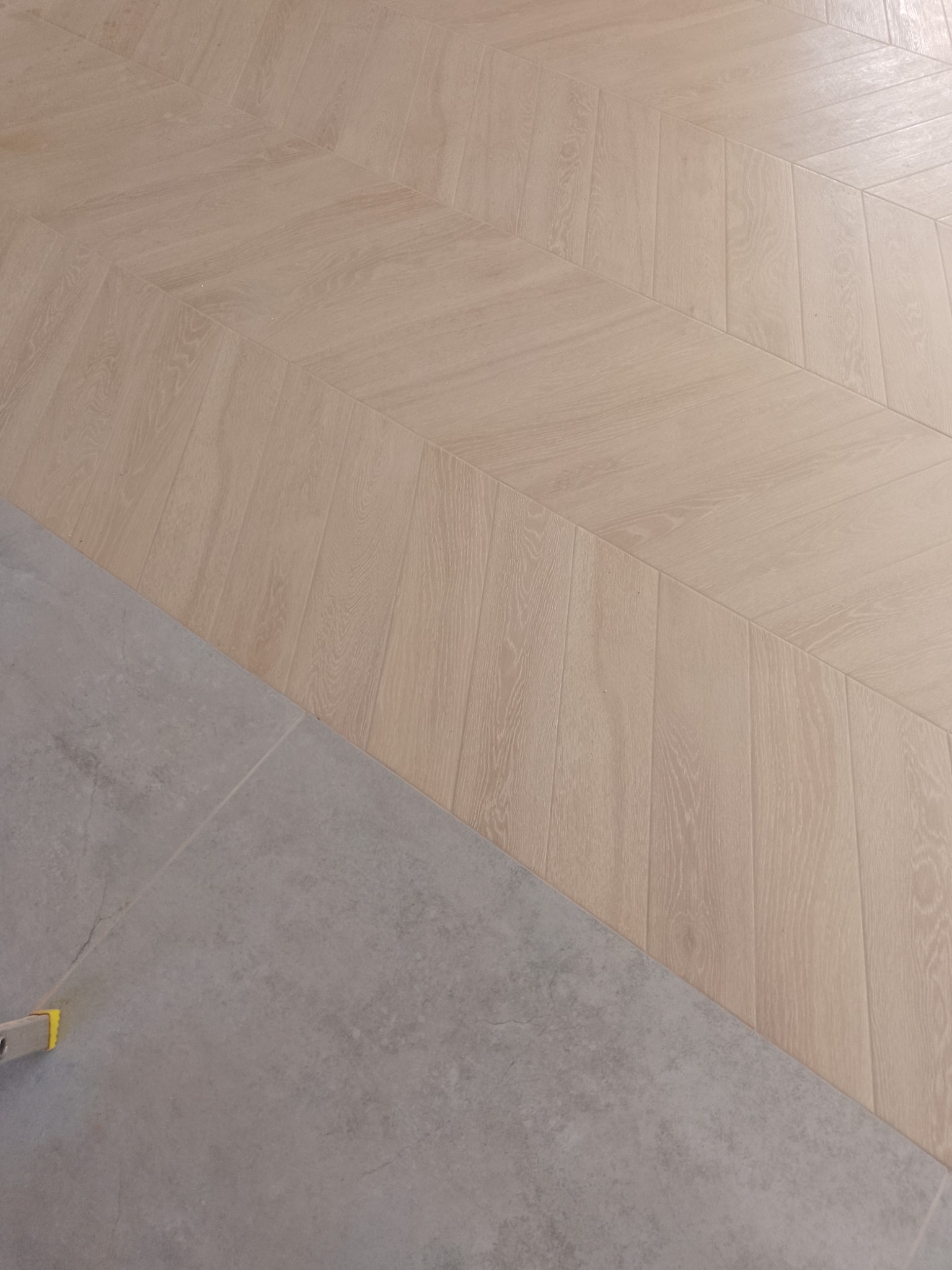 Jasne panele ułożone we wzór jodełki francuskiej, graniczące z szarą, matową płytką. Widoczny fragment poziomicy. Kontrast materiałów i faktur.