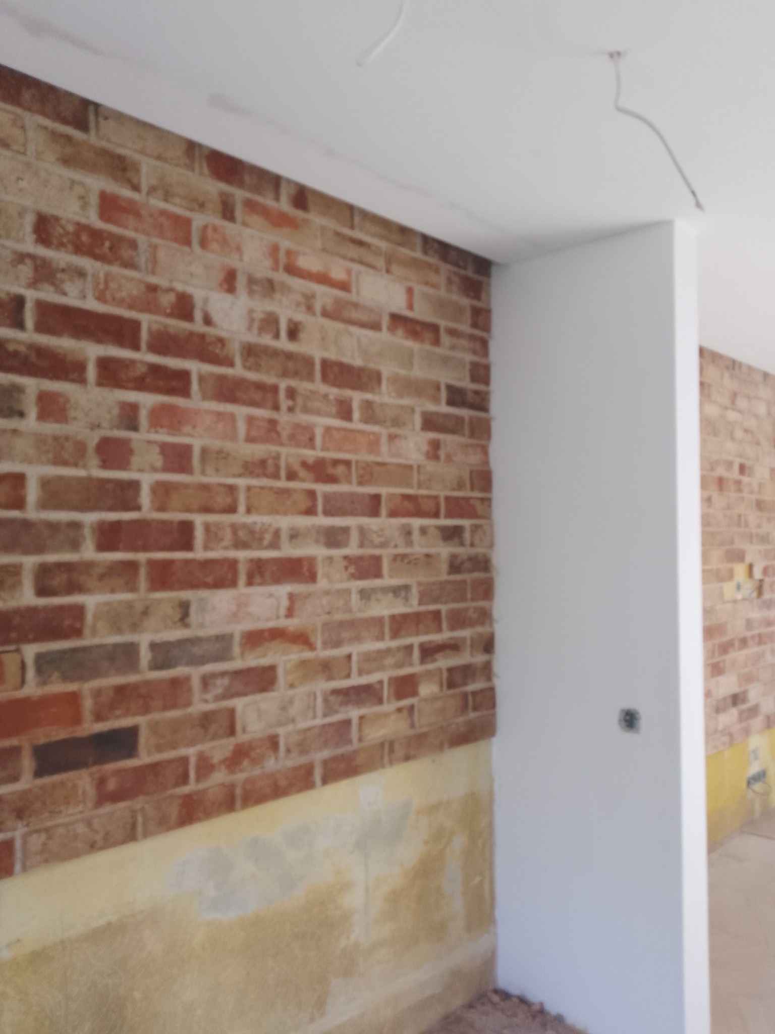 Ściana z cegły dekoracyjnej w pomieszczeniu w trakcie wykańczania, widoczny fragment surowego tynku i wystające przewody. Cegła w odcieniach czerwieni i brązu.