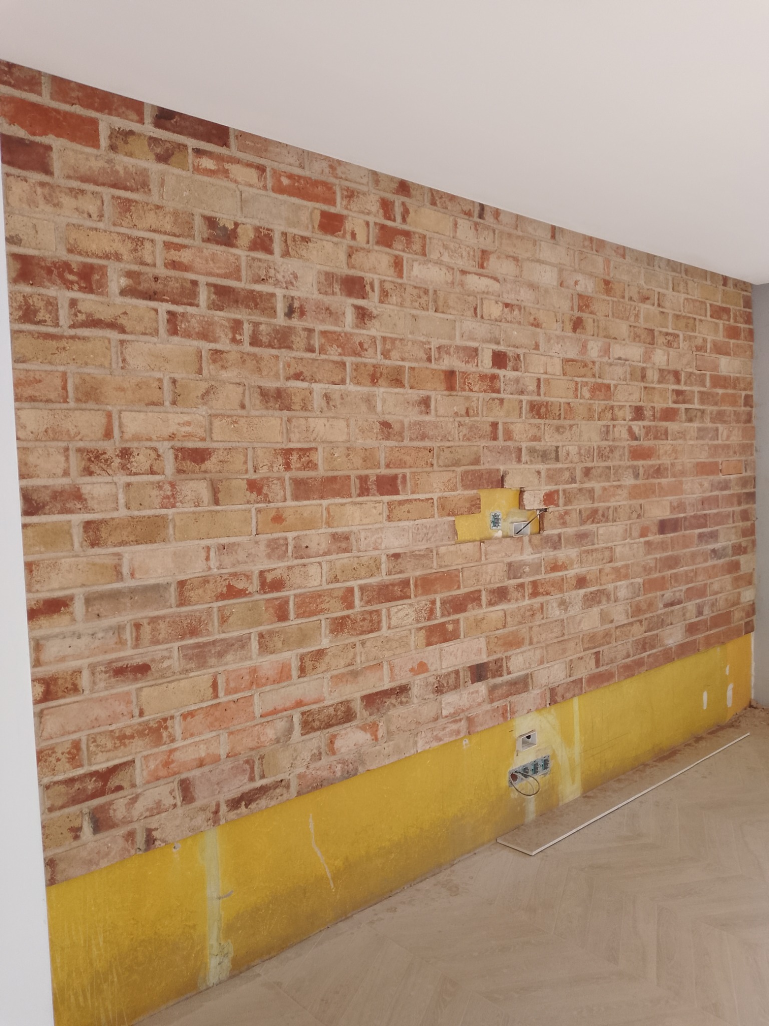 Ściana z cegły dekoracyjnej w trakcie montażu, zabezpieczona folią. Widoczne gniazdka elektryczne i fragment podłogi. Ujęcie z boku.