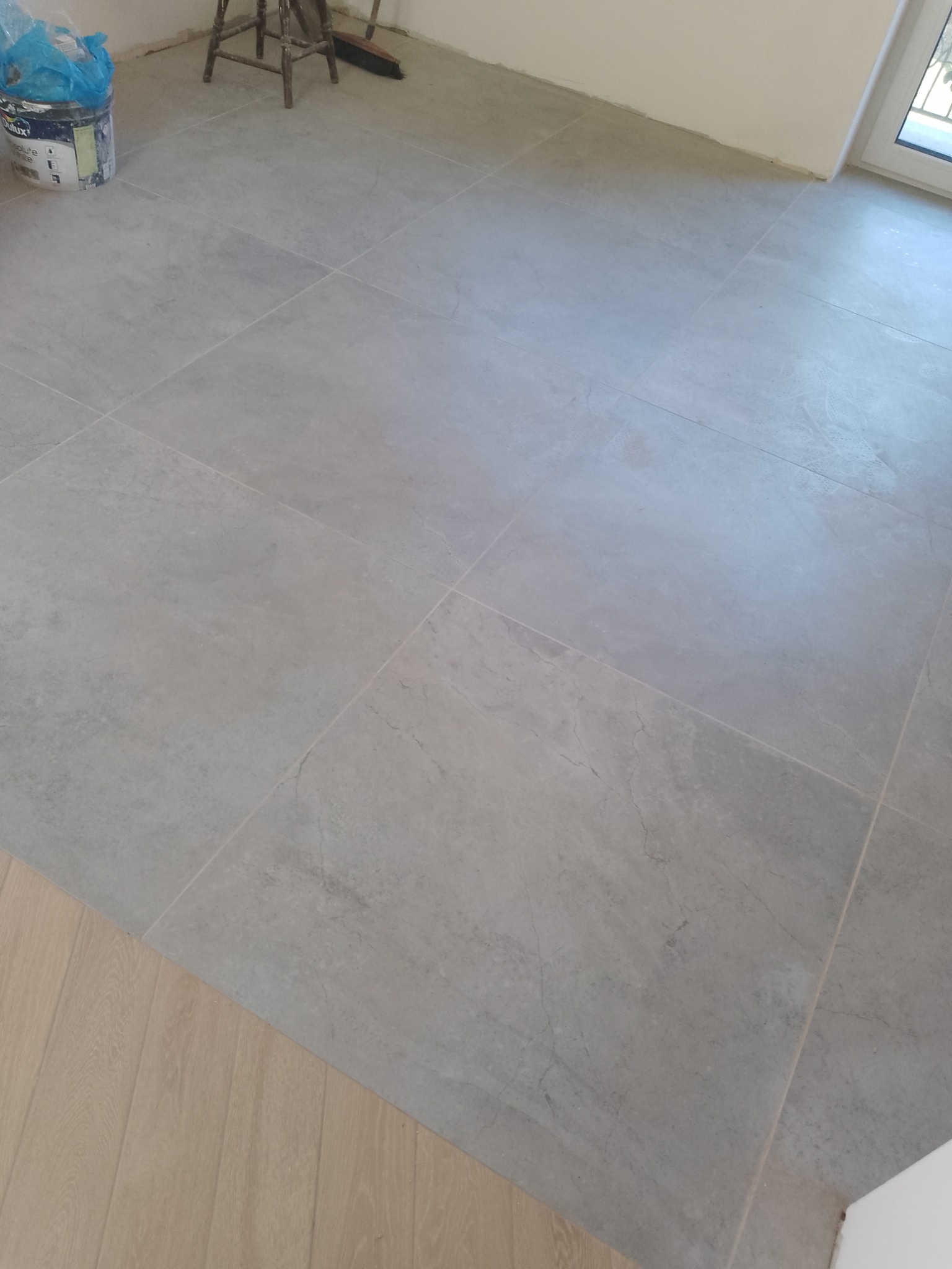 Ułożone szare płytki podłogowe w nowym pomieszczeniu, widoczny fragment drewnianej podłogi, wiadro i stołek w tle. Minimalistyczne wykończenie wnętrza.