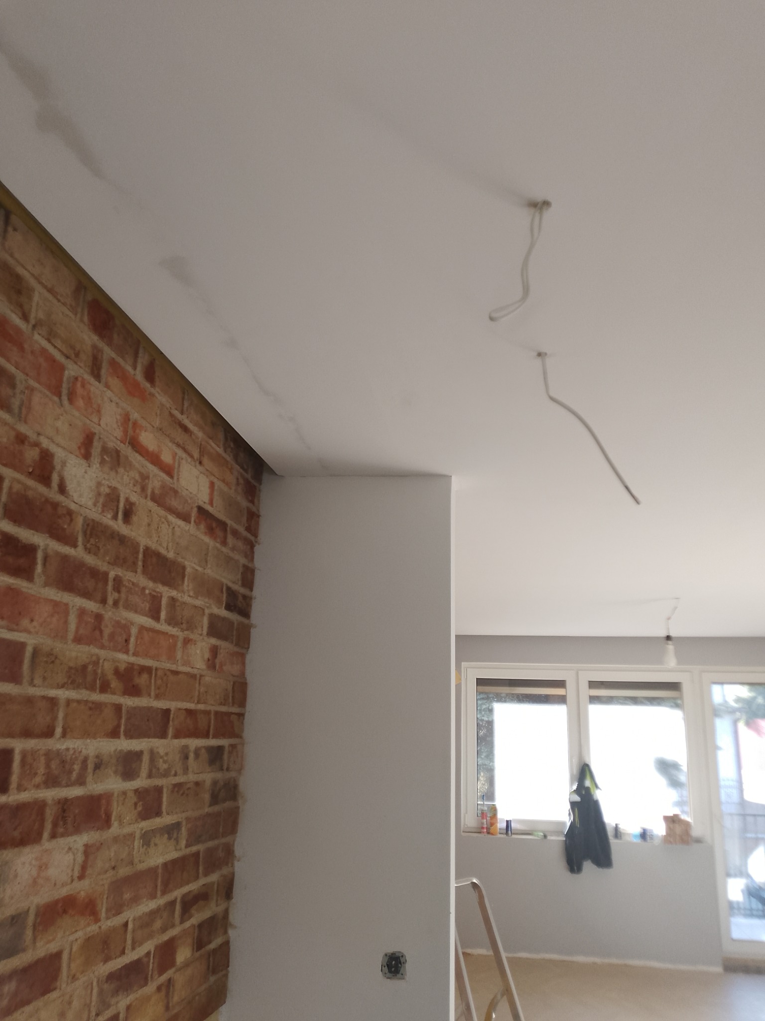 Wnętrze w trakcie prac wykończeniowych: ceglana ściana kontrastuje z pomalowanym na biało sufitem, widoczne kable oświetleniowe i drabina. Częściowo odnowione wnętrze.