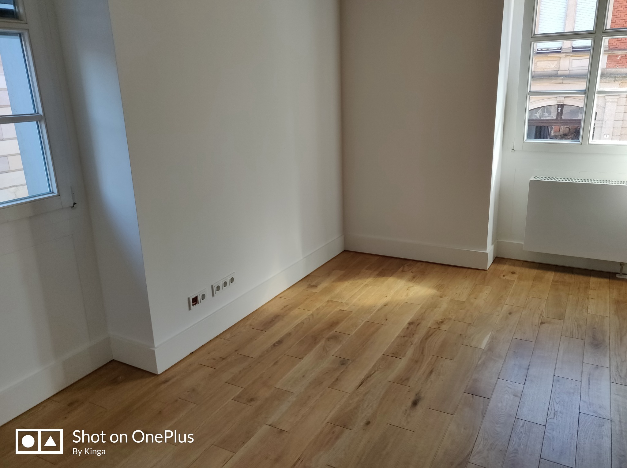 Jasne panele podłogowe w nowym, odmalowanym pokoju z białymi ścianami i listwami przypodłogowymi. Widoczne gniazdka elektryczne i okno z widokiem na budynek.