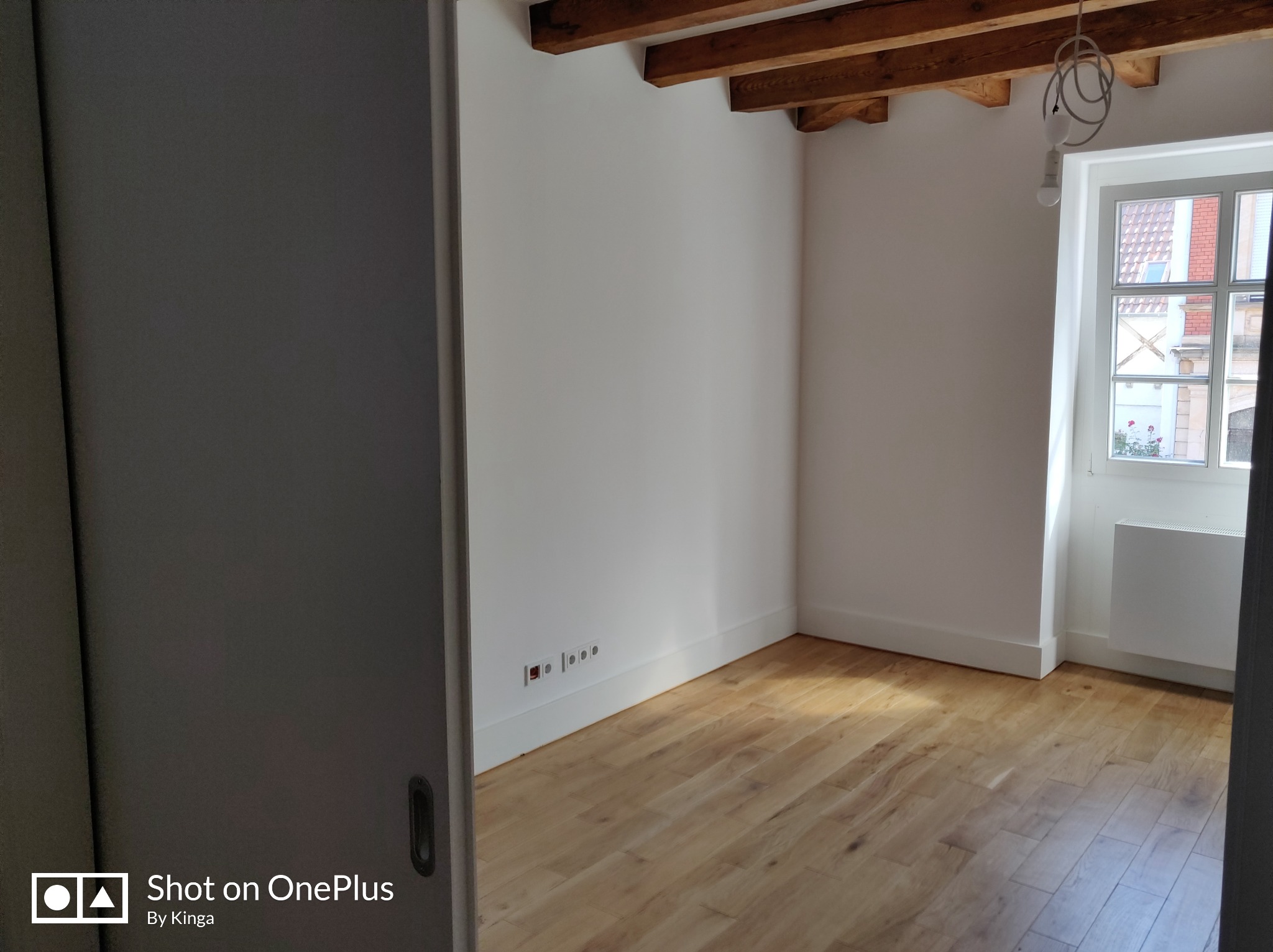 Jasne, odświeżone wnętrze z drewnianą podłogą, białymi ścianami i widocznymi belkami stropowymi. Okno z widokiem na ceglany budynek w tle. Minimalistyczny design.