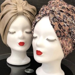 Antonina Melnychuk - Dwa manekiny z turbanami: beżowy, gładki i wzorzysty, cekinowy z motywem panterki. Czerwone usta i sztuczne rzęsy. Eleganckie nakrycia głowy na ekspozycji.
