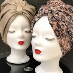 Dwa manekiny z turbanami: beżowy, gładki i wzorzysty, cekinowy z motywem panterki. Czerwone usta i sztuczne rzęsy. Eleganckie nakrycia głowy na ekspozycji.