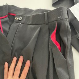 Antonina Melnychuk - Czarne spodnie z zakładkami i czerwoną podszewką kieszeni, w trakcie szycia, z fragmentem materiału w tle. Widoczna ręka na spodniach.