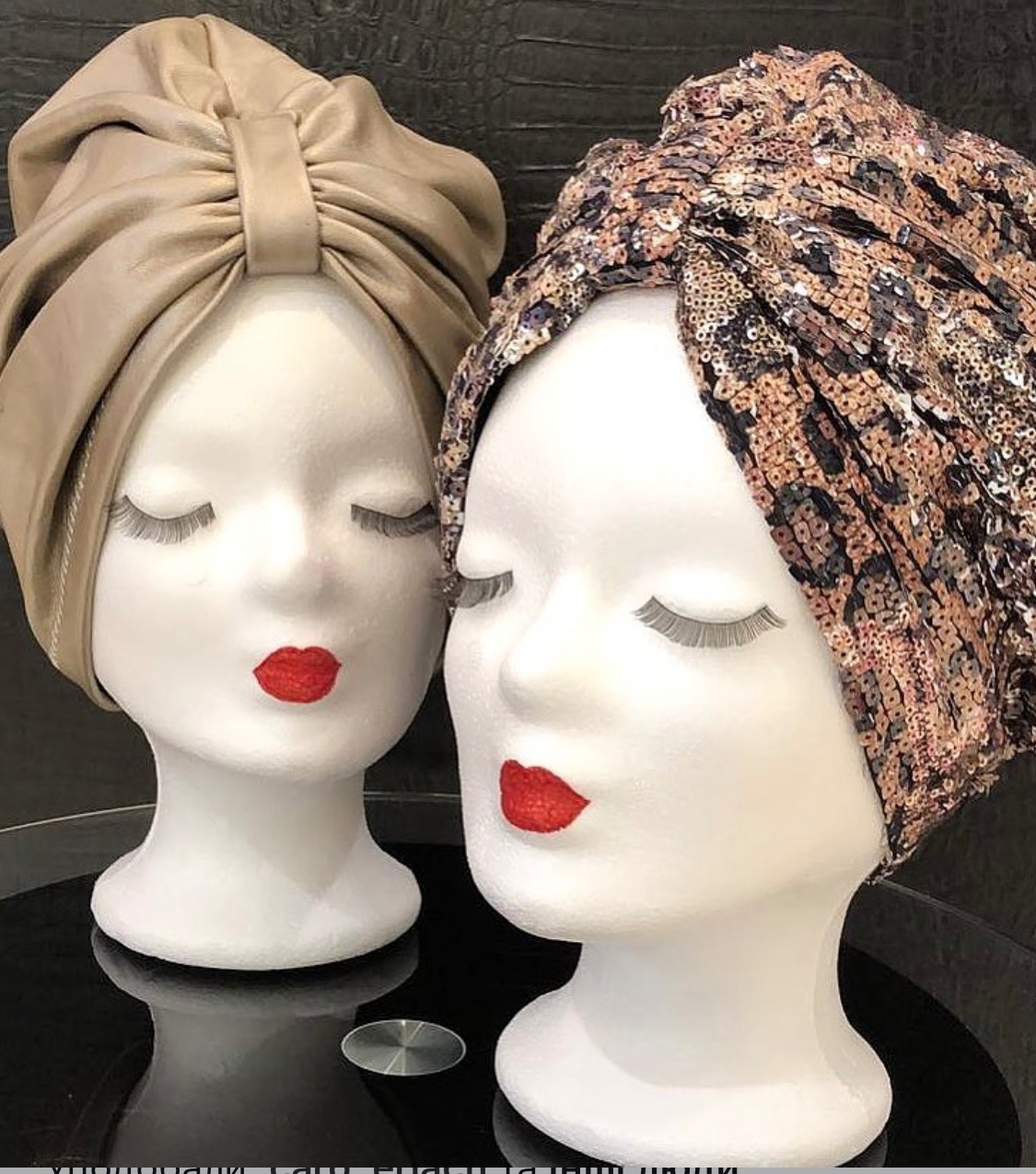 Dwa manekiny z turbanami: beżowy, gładki i wzorzysty, cekinowy z motywem panterki. Czerwone usta i sztuczne rzęsy. Eleganckie nakrycia głowy na ekspozycji.