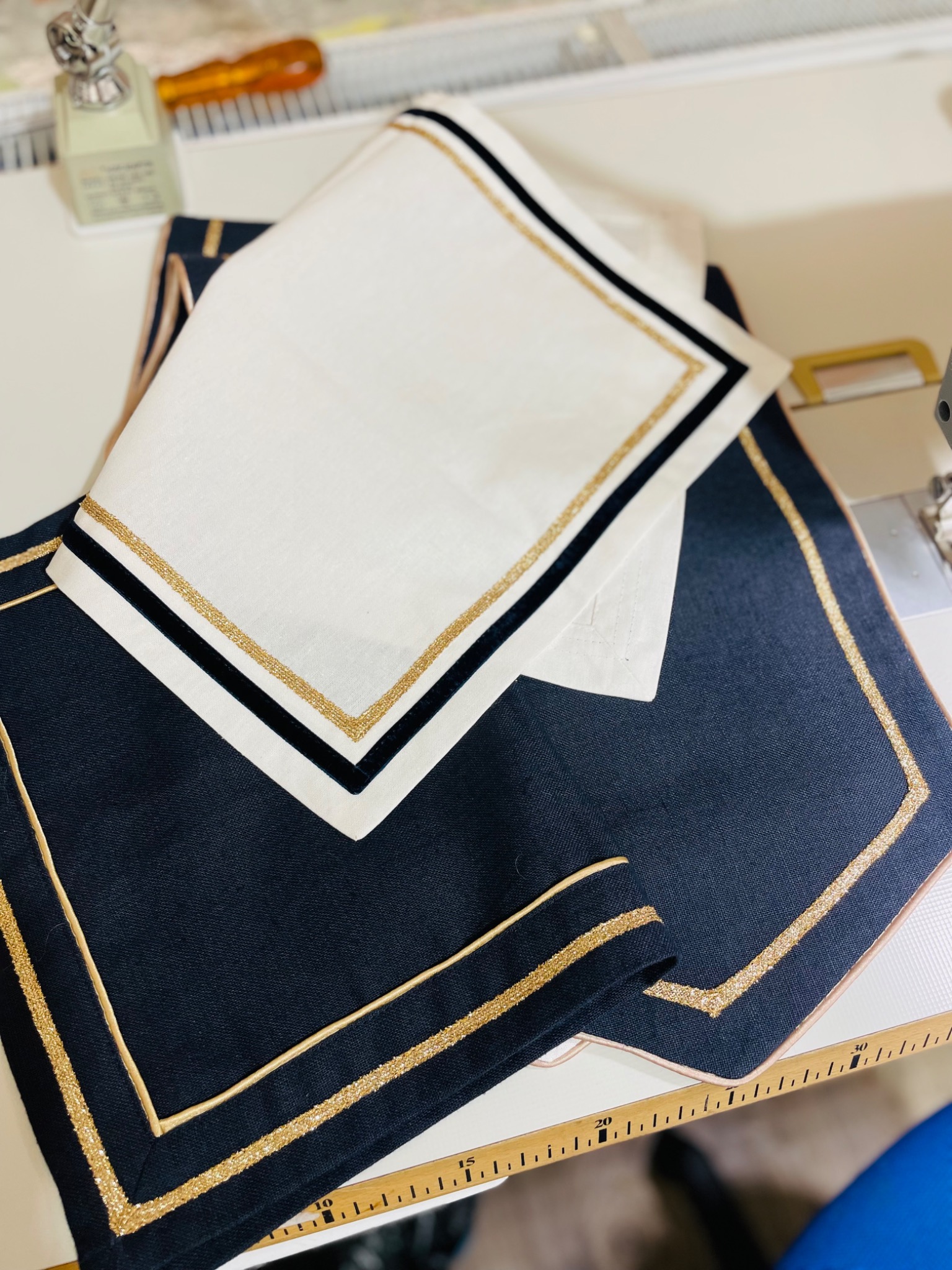 Eleganckie, ręcznie szyte podkładki na stół z granatowego i białego lnu, zdobione złotą lamówką i czarnym pasem, leżące na stole obok maszyny do szycia.