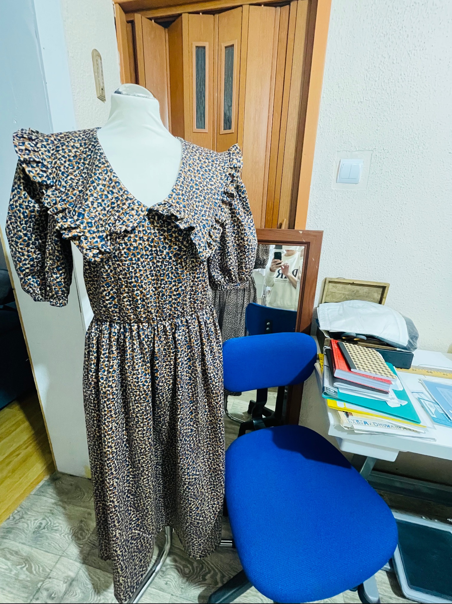 Sukienka w brązowo-niebieski wzór na manekinie, z falbankami przy dekolcie i krótkimi rękawami, stojąca w pomieszczeniu z biurkiem i krzesłem.
