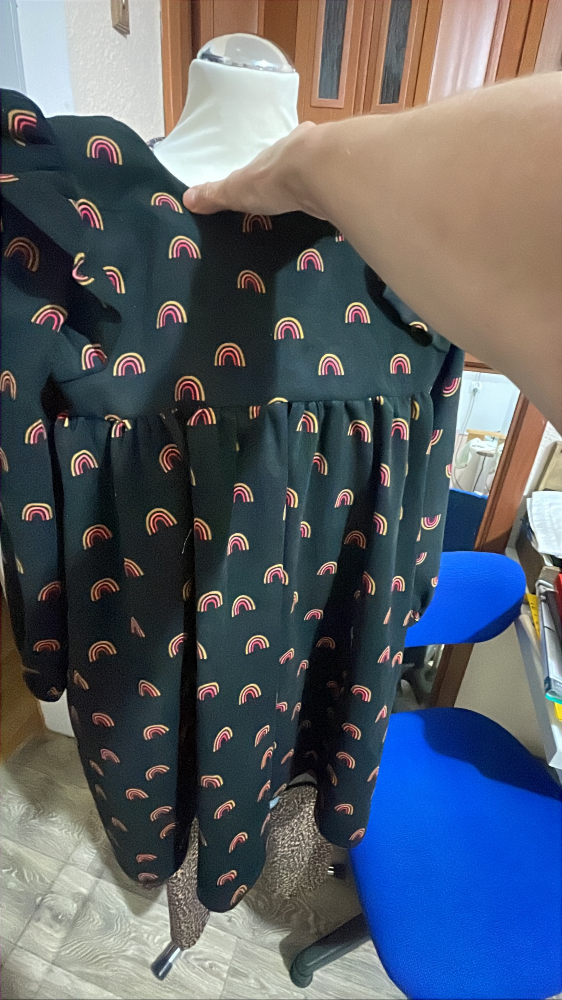 Czarna sukienka w tęcze na manekinie, trzymana przez rękę. Widoczne detale wzoru i krój sukienki. W tle biurowe krzesło i meble.