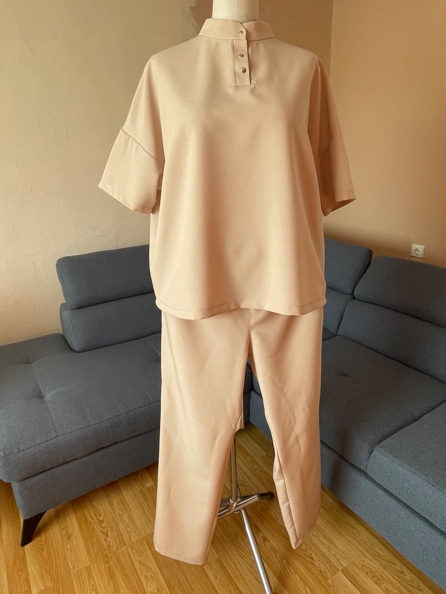 Beżowy, damski komplet dresowy z krótkim rękawem na manekinie, na tle szarej sofy. Bluzka z trzema guzikami przy szyi, spodnie proste.