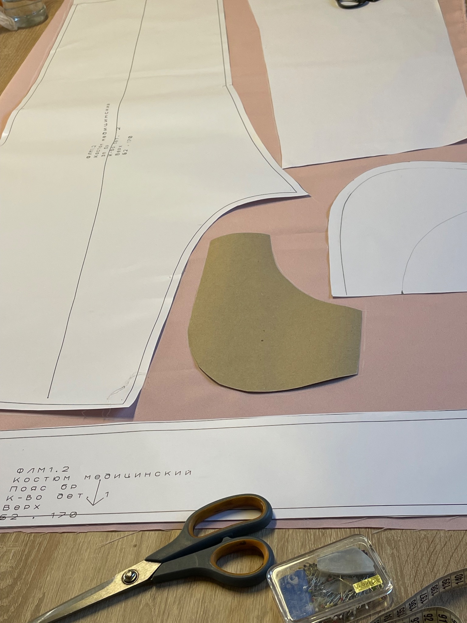 Rozłożone wykroje odzieży medycznej na różowym materiale, obok leżą nożyczki, szpilki i miarka krawiecka. Widoczne napisy w języku rosyjskim na wykrojach.