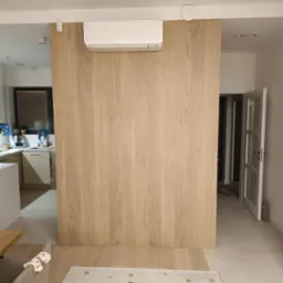 Nowoczesna zabudowa ściany z paneli drewnianych w jasnym odcieniu, zintegrowana z klimatyzacją. Minimalistyczny design w otwartym wnętrzu z widoczną kuchnią.