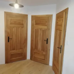 Trzy drewniane drzwi wewnętrzne z czarnymi klamkami, oświetlone lampą wiszącą, ukazane z perspektywy podłogi, eksponują naturalną fakturę drewna.