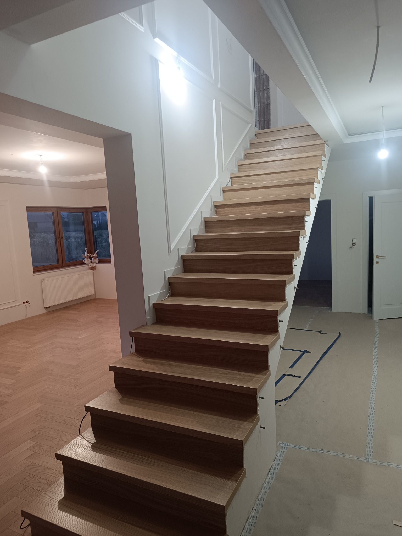 Jasne, nowoczesne schody z drewnianymi stopniami w nowym domu. Widok na salon z parkietem w jodełkę i białe ściany z dekoracyjnymi listwami.