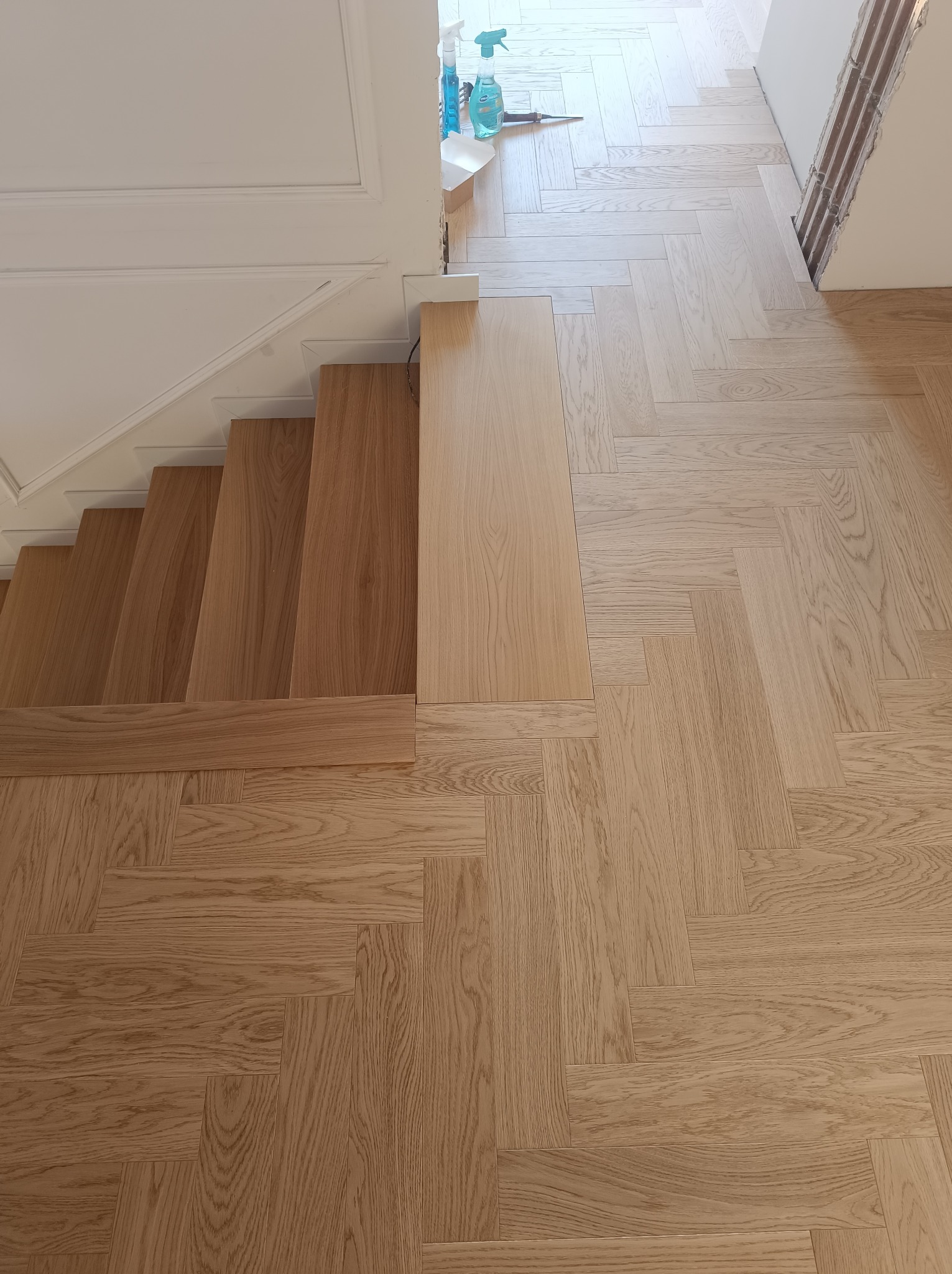 Ukończone schody i parkiet w jodełkę z jasnego drewna, widok z góry. Fragment białej ściany i środki czystości w tle. Detal wykończenia wnętrza.
