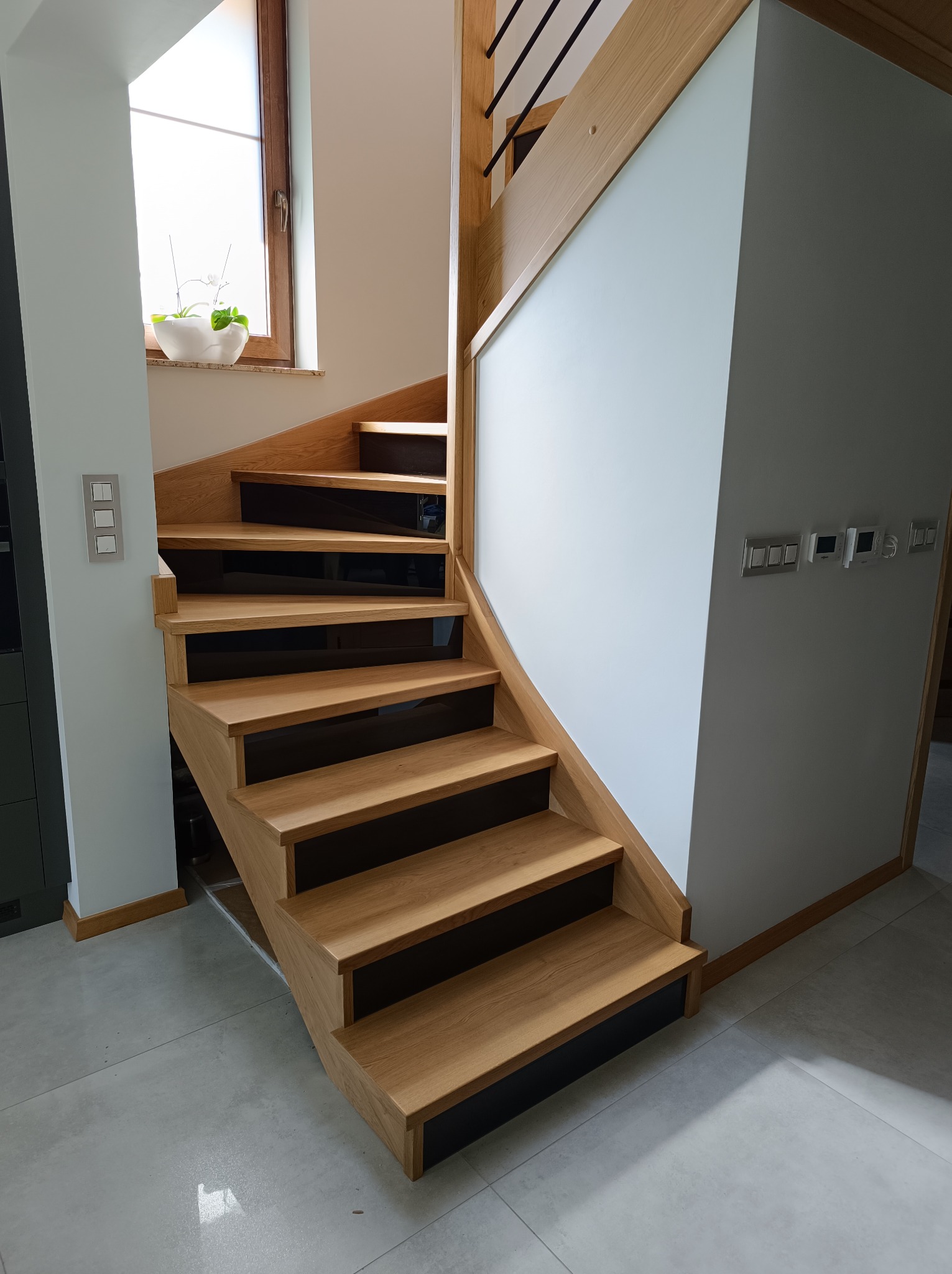 Nowoczesne schody z drewna dębowego z czarnymi podstopnicami, oświetlone naturalnym światłem z okna. Minimalistyczny design, jasne ściany, szare płytki podłogowe.