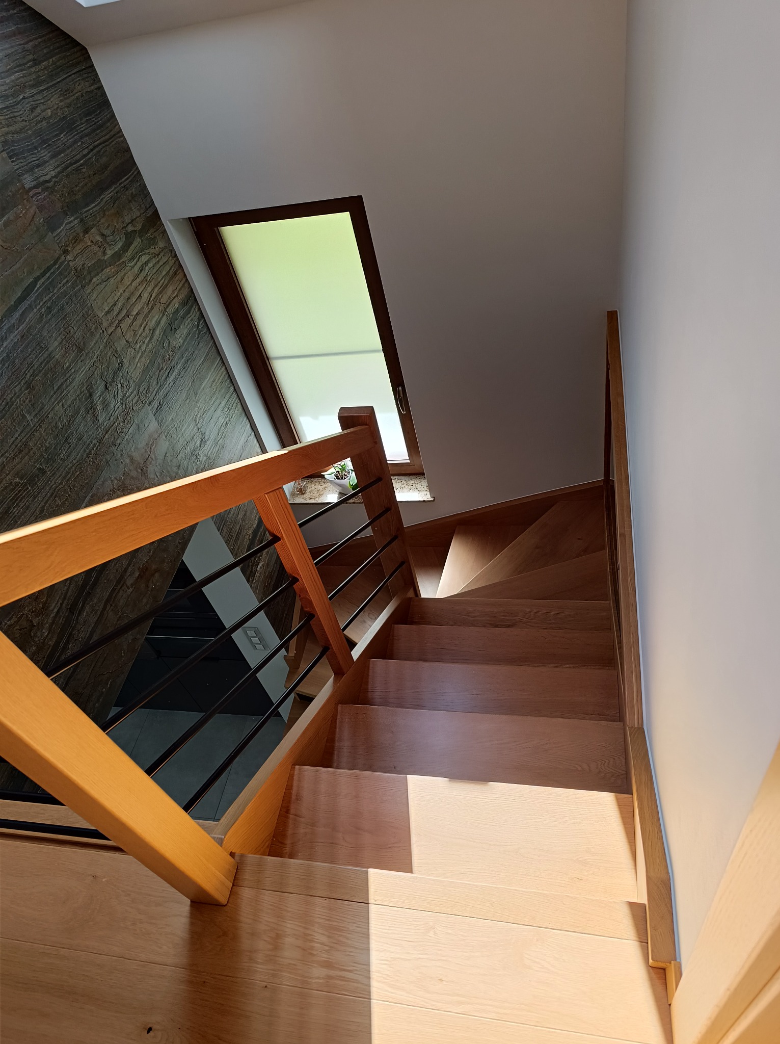 Jasne, drewniane schody z metalową balustradą w nowoczesnym wnętrzu. Widok z góry, naturalne światło z okna. Ściana z dekoracyjnym kamieniem.