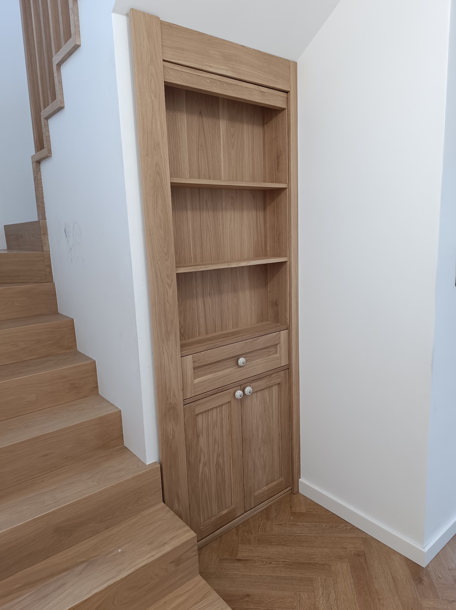 Drewniany regał wnękowy z szufladą i szafką, wkomponowany w przestrzeń pod schodami. Widoczne drewniane schody i parkiet w jodełkę. Jasne wnętrze.