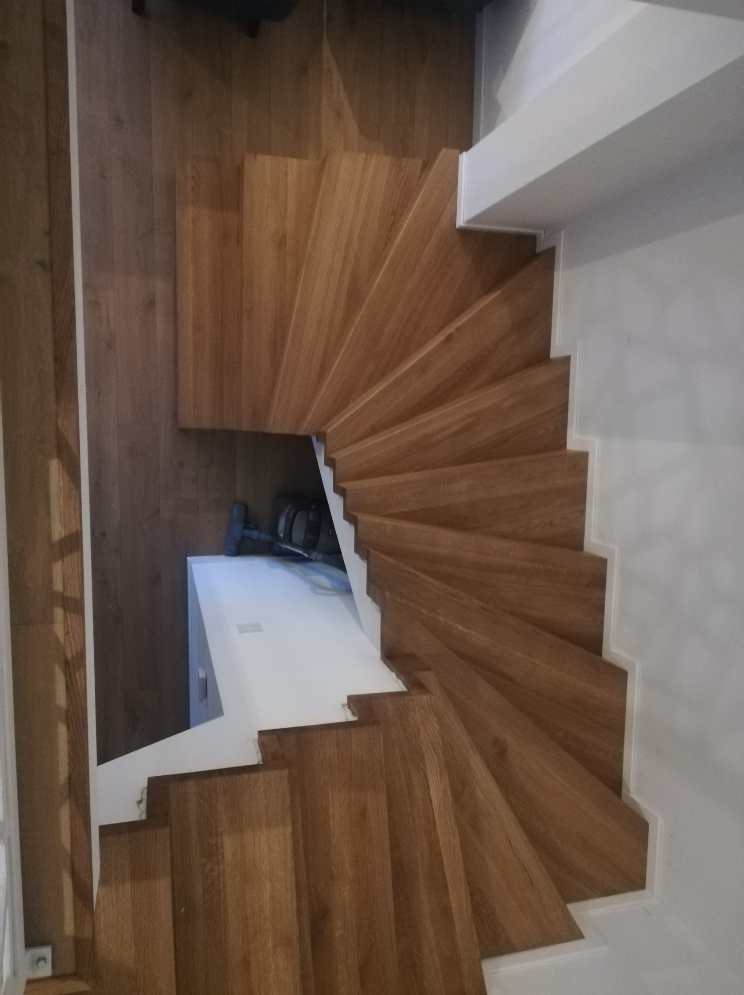 Widok z góry na kręte schody z drewna dębowego, z białymi ścianami i podstopnicami. Pod schodami widoczny odkurzacz. Parkiet na podłodze.