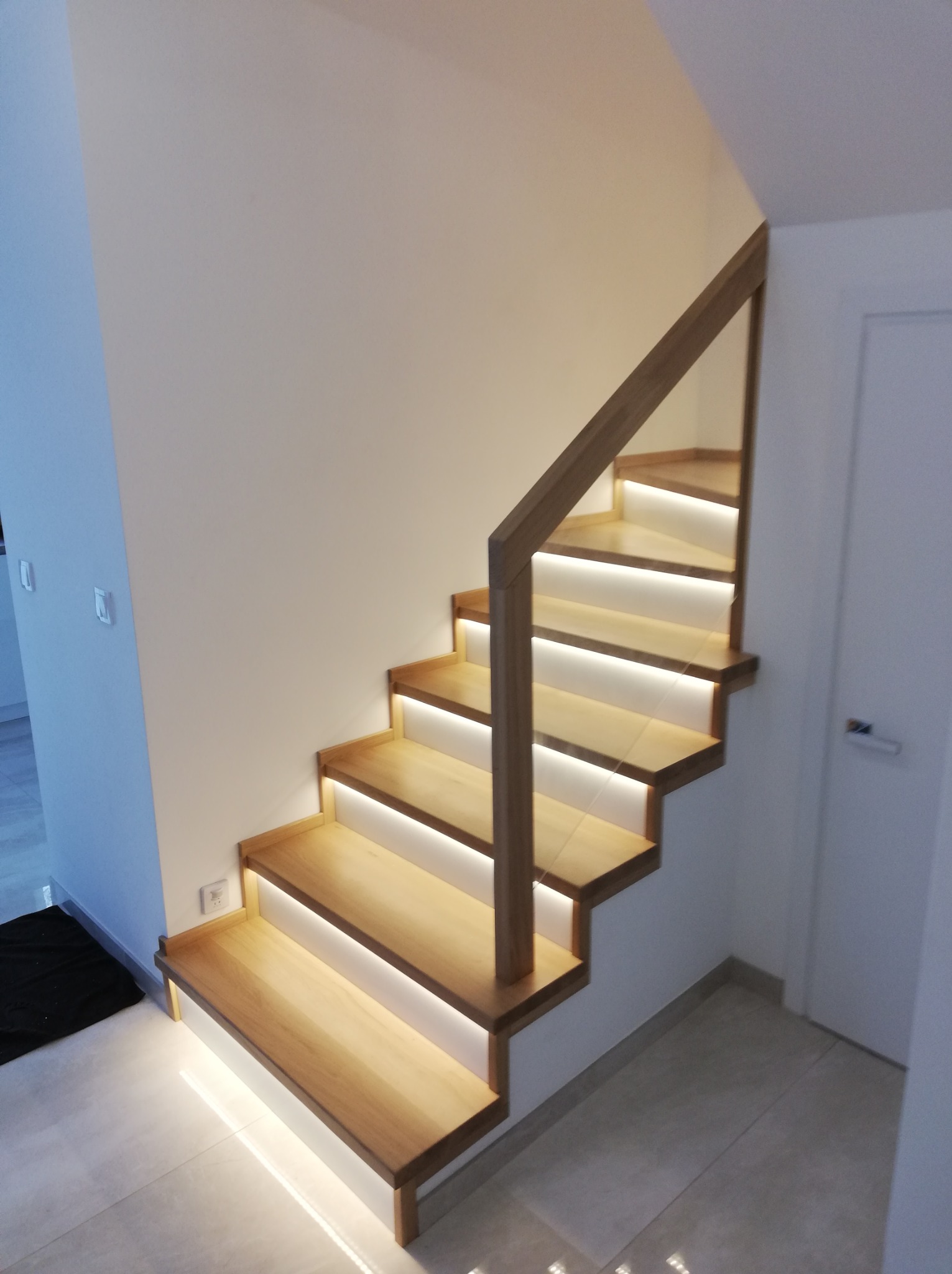 Nowoczesne schody z jasnego drewna z podświetleniem LED, widoczne z dołu. Minimalistyczna balustrada i białe ściany dopełniają aranżację wnętrza.