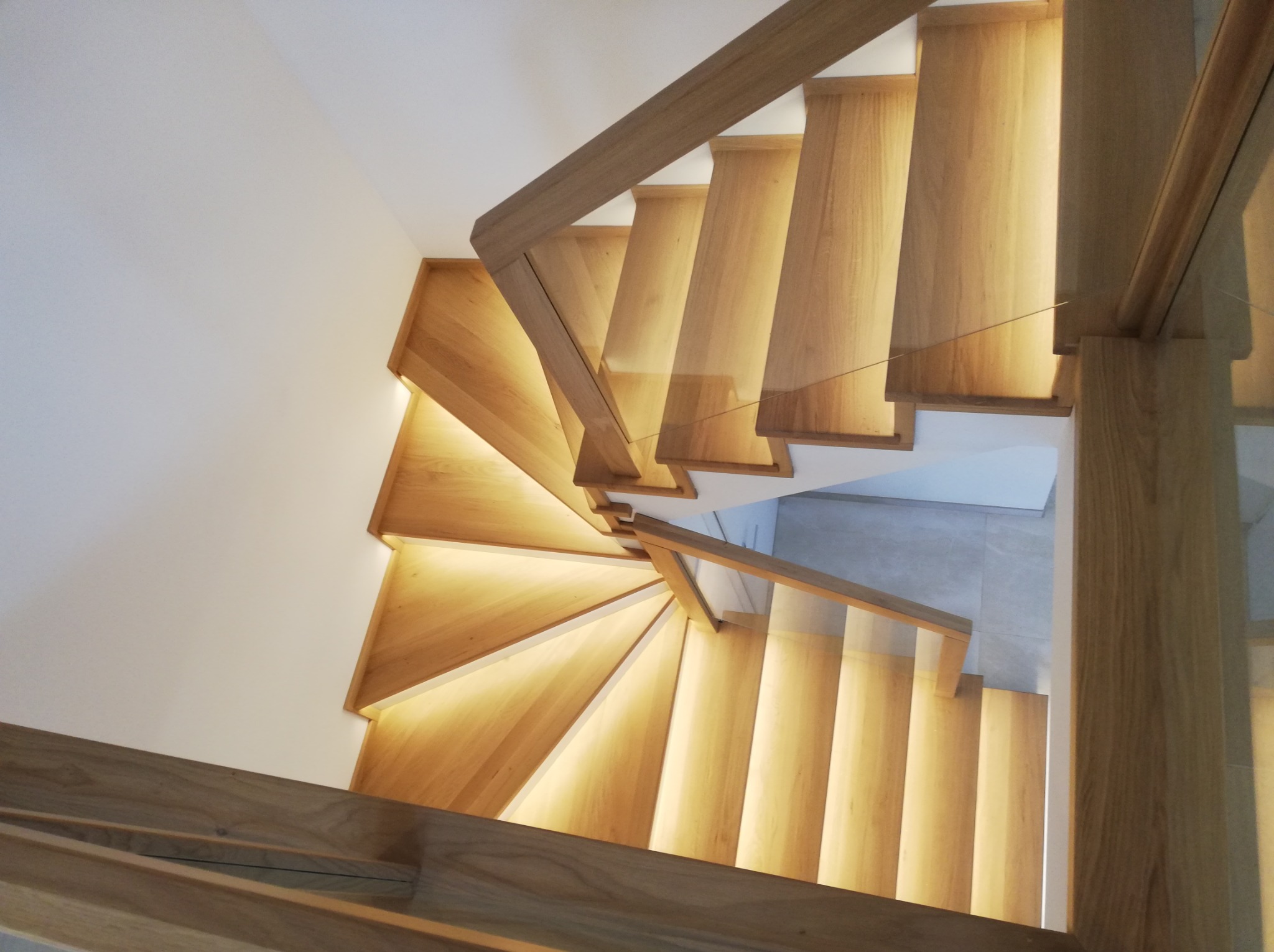 Spiralne schody z jasnego drewna z podświetleniem LED, widok z góry, z drewnianą balustradą i elementami szklanymi. Minimalistyczny design i nowoczesne oświetlenie.