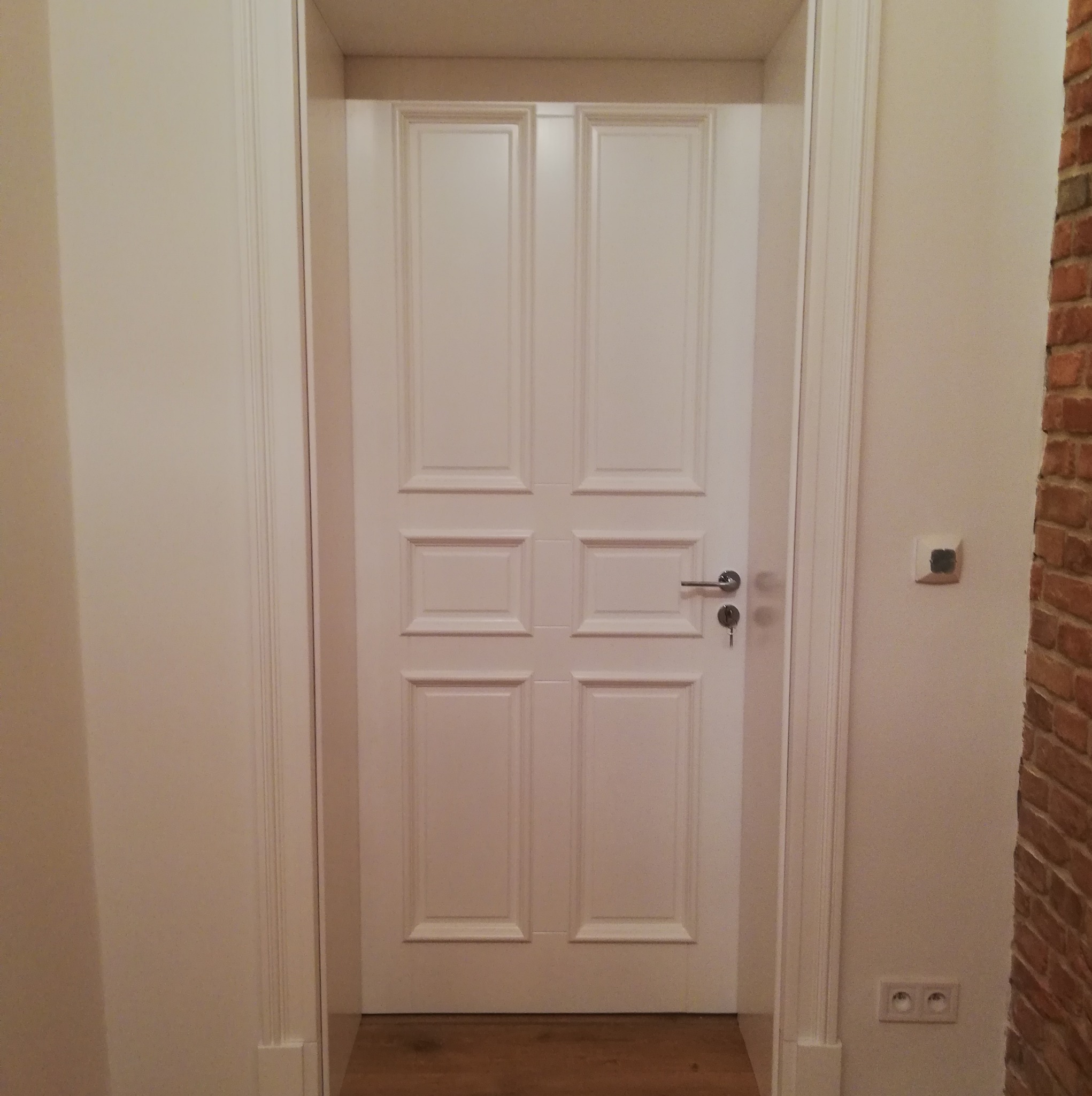 Białe, panelowe drzwi wewnętrzne z klamką i kluczem, widoczne w korytarzu z ceglaną ścianą w tle. Klasyczny design, świeżo po montażu.